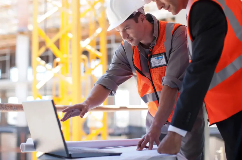 Two engineers in safety vests reviewing project details on a laptop
